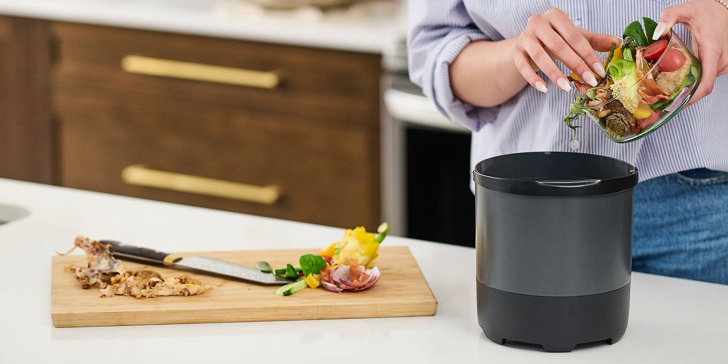 A person in a blue shirt pouring a variety of food scraps from a glass bowl into a dark gray FoodCycler bucket. On the kitchen counter next to the bucket, there is a wooden cutting board with a knife and more vegetable trimmings.