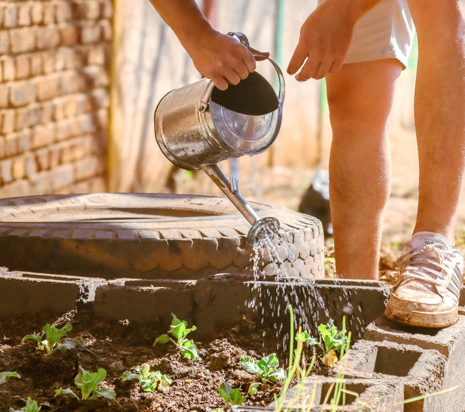 Feet of a person with a simple tin watering can, standing on repurposed tires and cinderblocks for a makeshift, low-cost garden