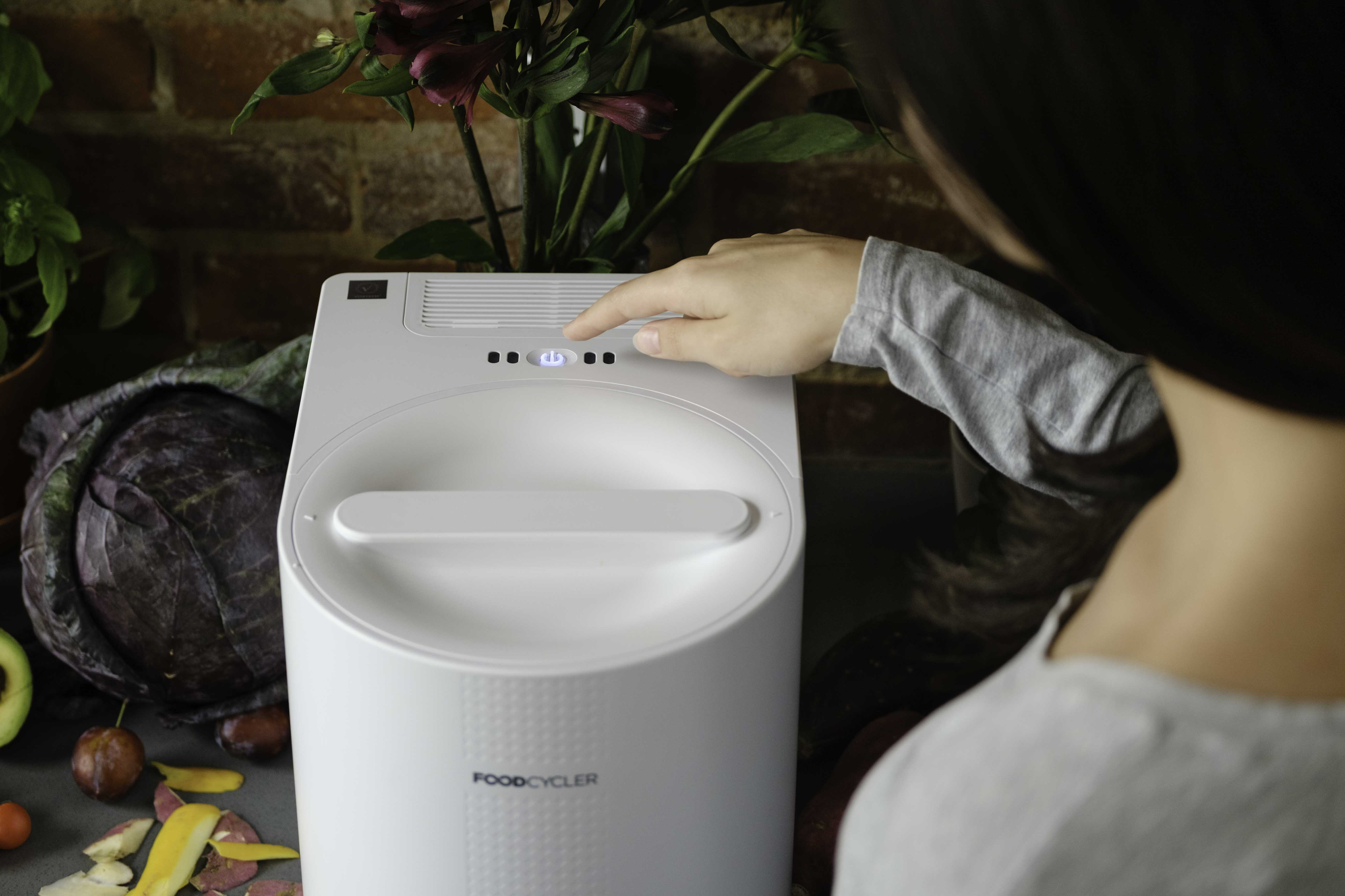 A person's hand presses the illuminated blue power button on top of the white FoodCycler Eco 3. The appliance is situated on a kitchen counter surrounded by a red cabbage, food scraps, and potted plants against a brick wall background.