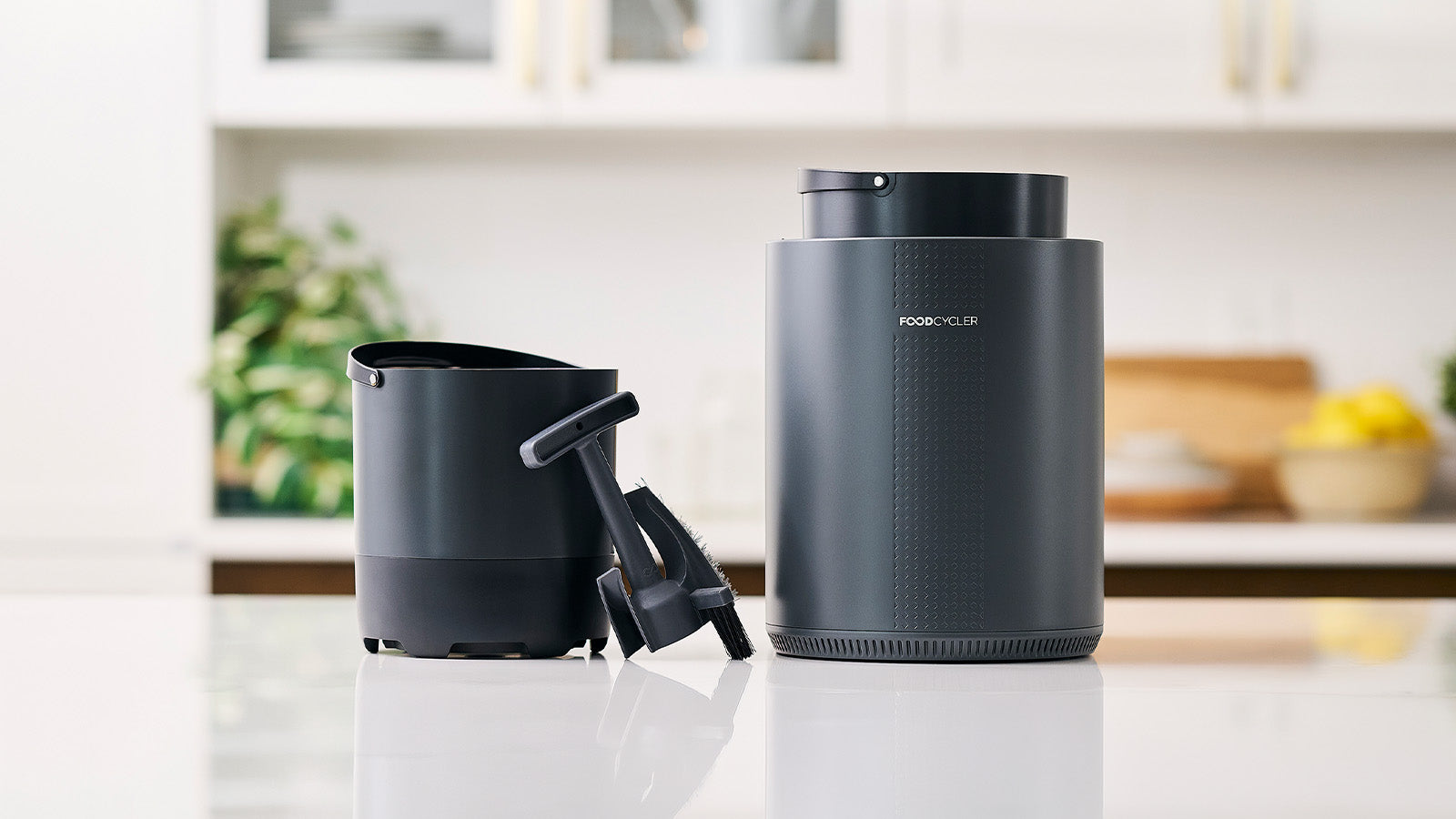 Le seau FoodCycler Eco avec la brosse InfinityBrush, posé à côté de l'unité de recyclage alimentaire Eco sur un plan de travail de cuisine