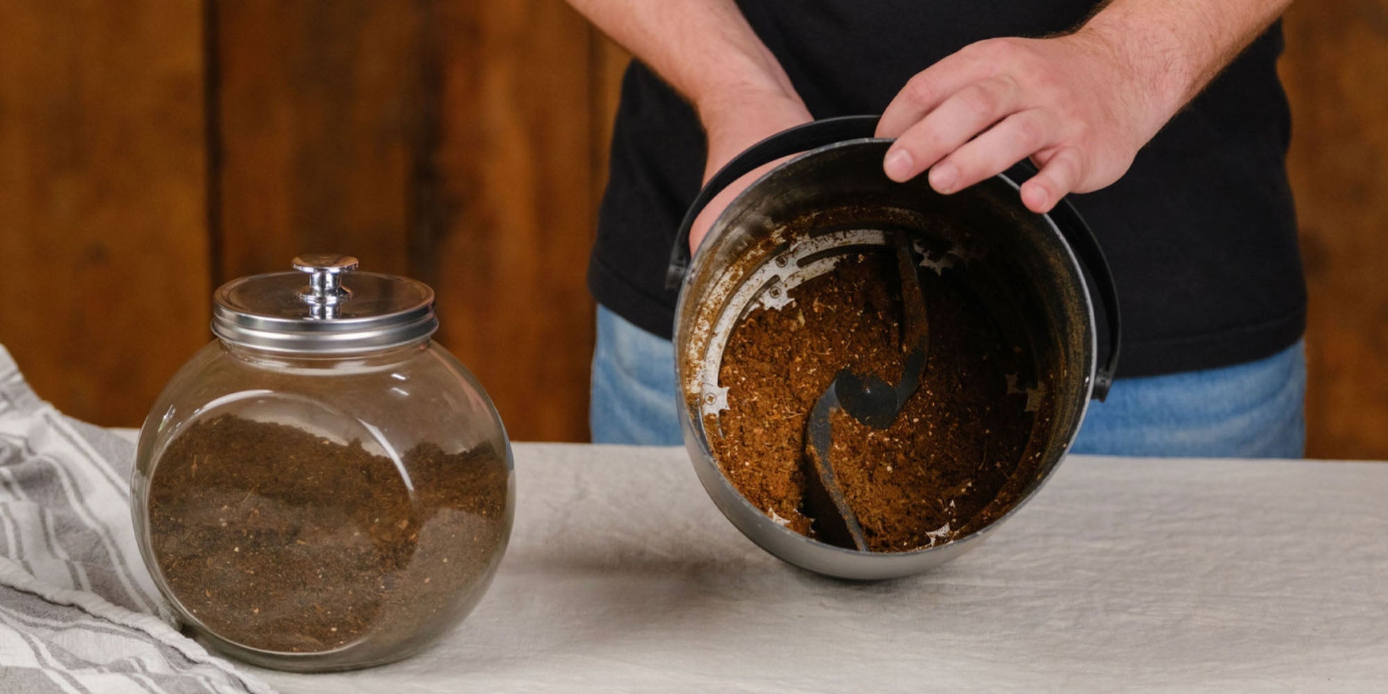 A person holds the inner bucket of a FoodCycler, showing the dark brown, processed organic matter inside. On the table next to them is a large glass jar filled with the same soil-like material.