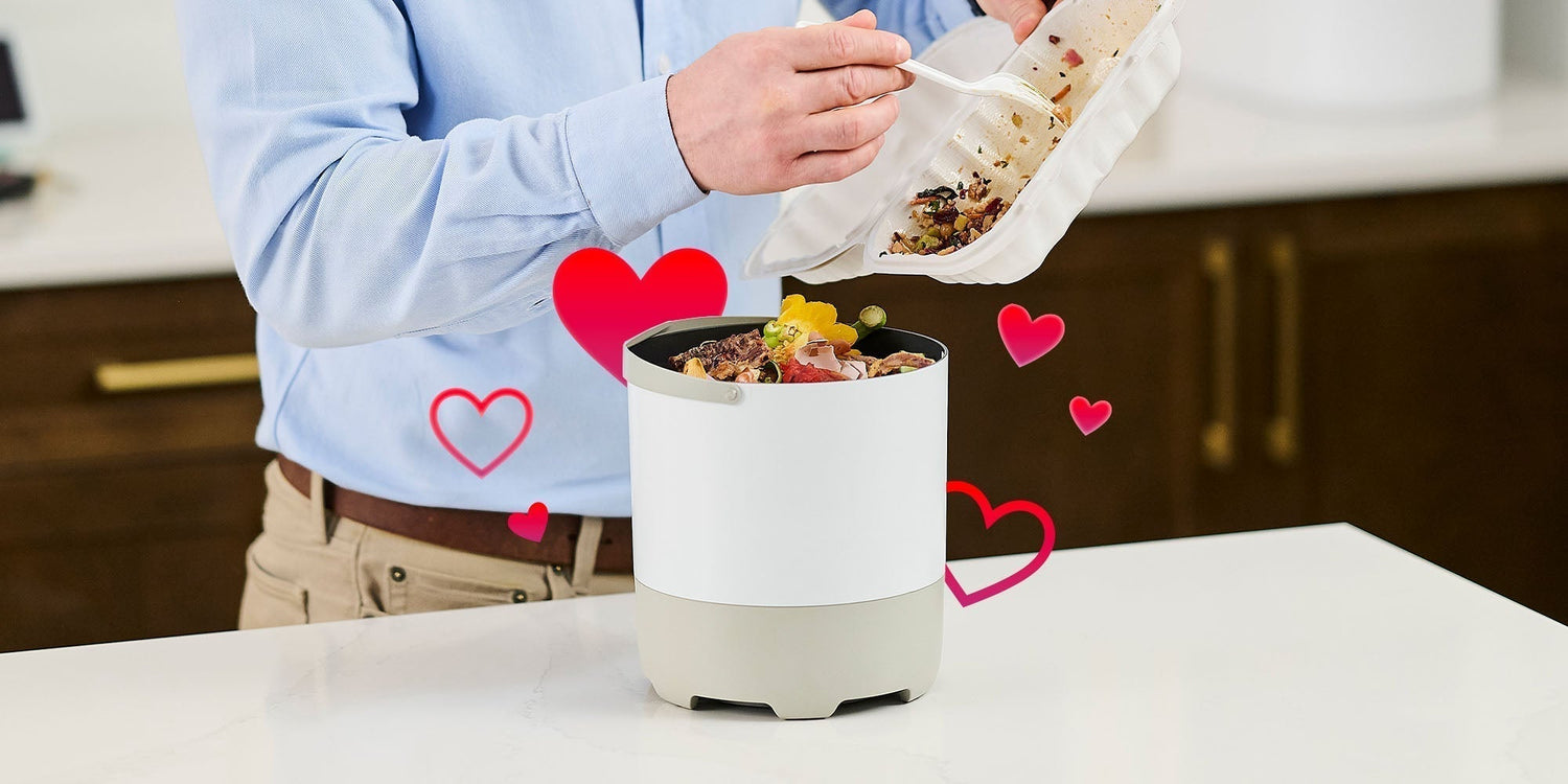 A person scraping food leftovers from a white takeout container into a countertop compost bucket, surrounded by floating red heart graphics.