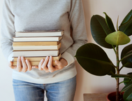 A close-up of a person in a gray sweater and blue jeans holding a heavy stack of five hardcover books. A large-leafed rubber plant is visible in the background to the right.