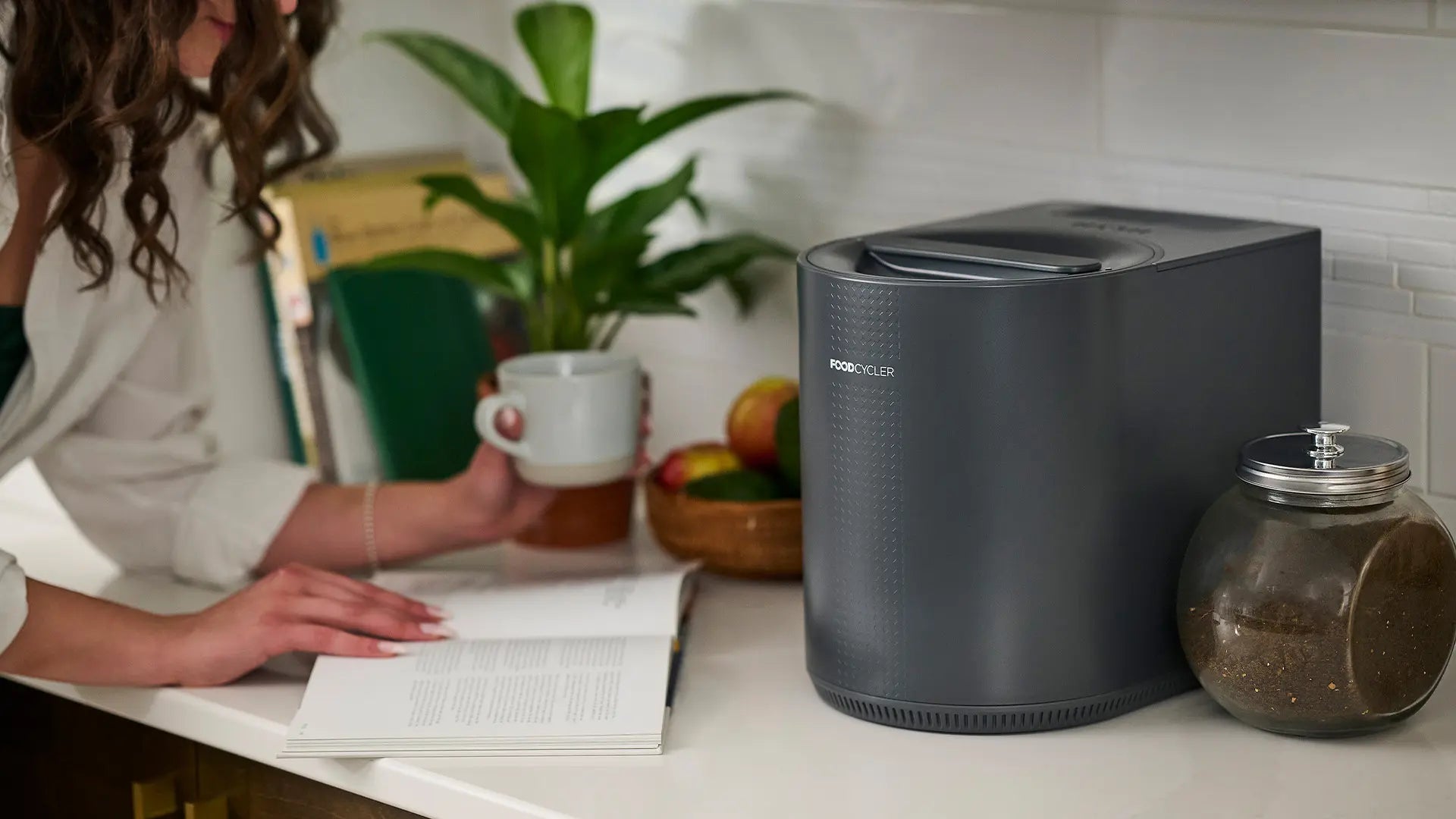 Person reading a book and drinking coffee on kitchen counter; gray FoodCycler Eco 3 next to them. 
