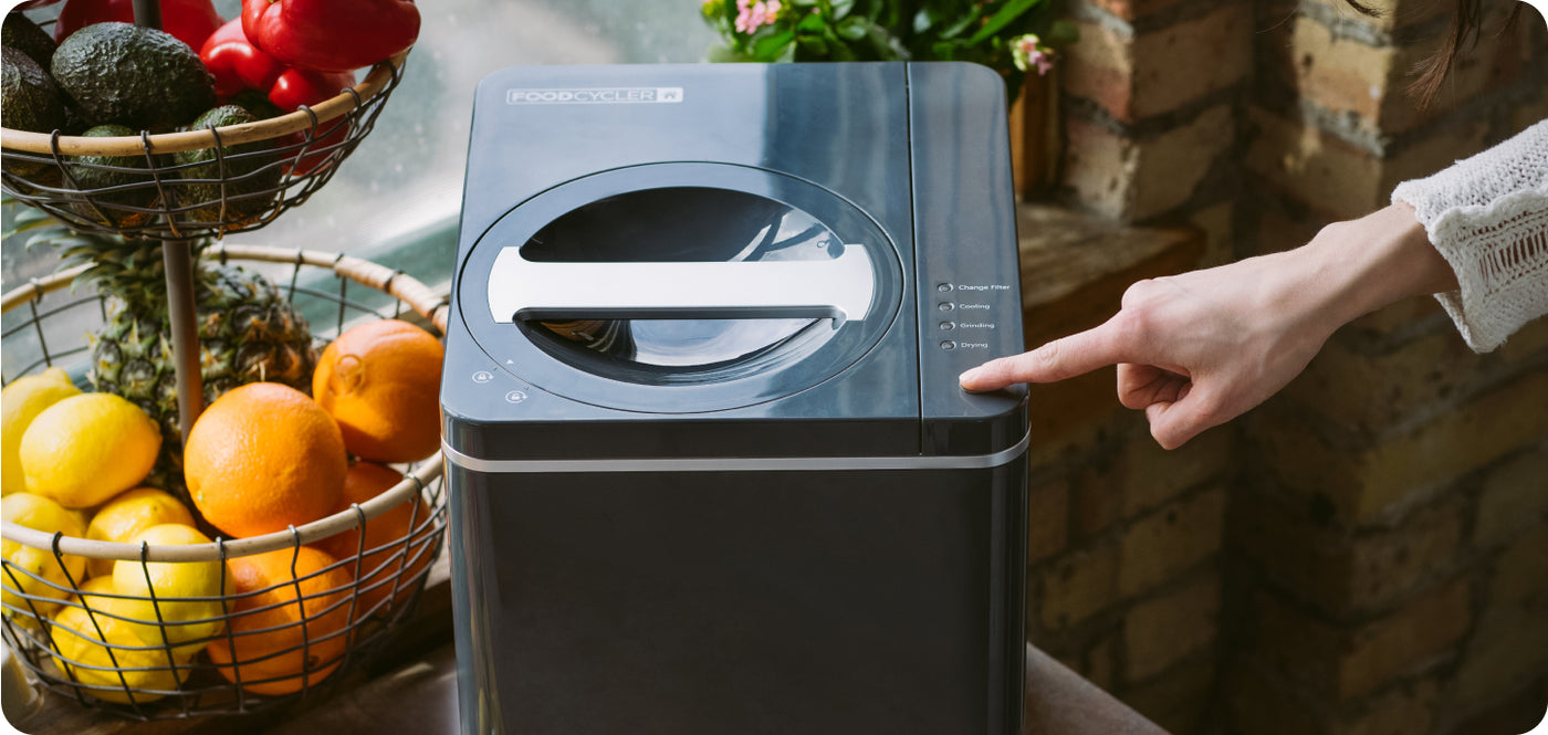 A hand pointing to the indicator lights on a FoodCycler FC-30 sitting on a kitchen counter beside a basket of fruit